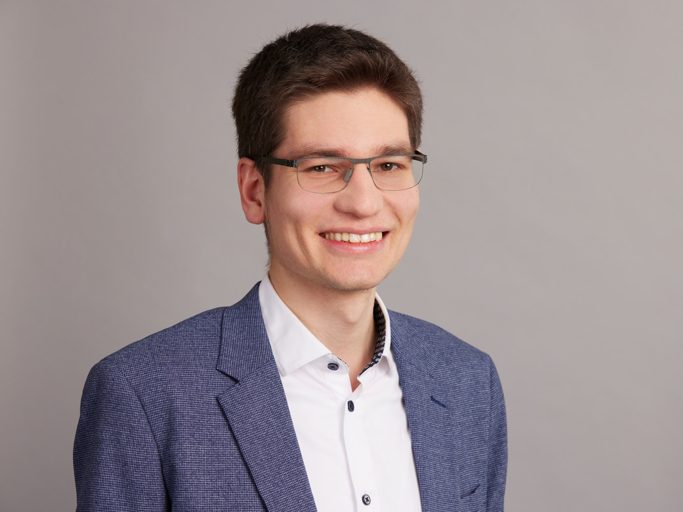 Photo of Wolfgang Bubich, a smiling younger man with short hair and eyeglasses. He is wearing a blue blazer and a white shirt.